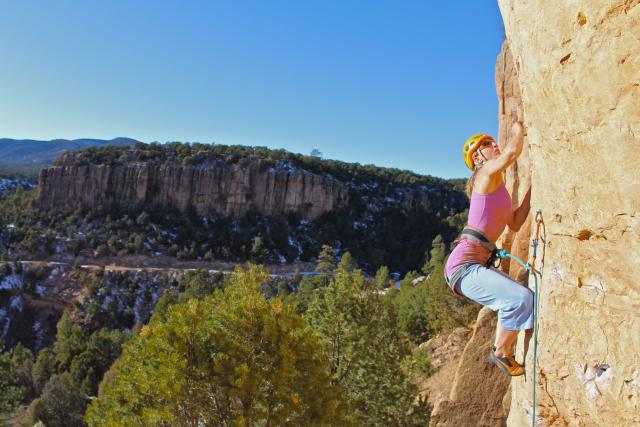 A blonde woman in the foreground wearing a helmet, sunglasses, tank top, capri pants, and climbing shoes is harnessed at the waist into the canyon wall as she ascends the right side of the image with mature evergreen trees below her. Cloudless blue sky, mountains, and another canyon wall are in the background peppered by evergreen shrubs and patches of snow.