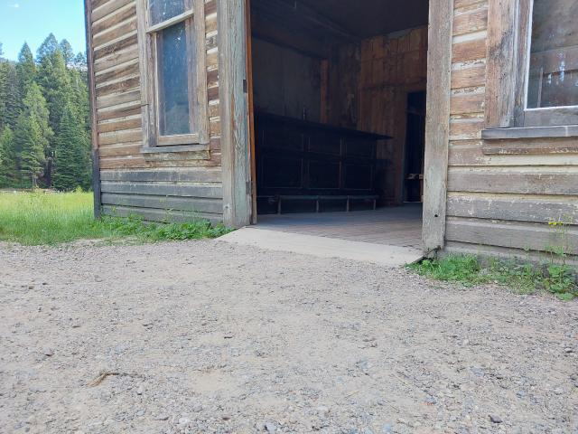 A photo showing a packed gravel surface that leads to a wide wooden threshold into a historic wooden building with a wide doorway.