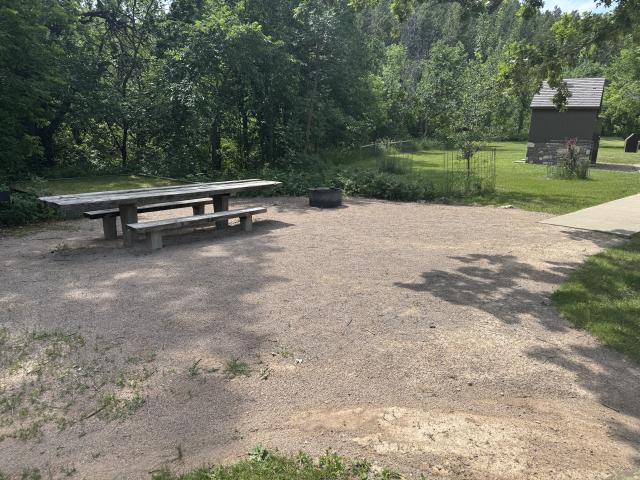 A photo showing a packed gravel campsite with a long table that has extended ends to accommodate visitors using a wheelchair. A metal firepit is next to the table. A sidewalk goes out of frame towards the vault toilet building in the background. 