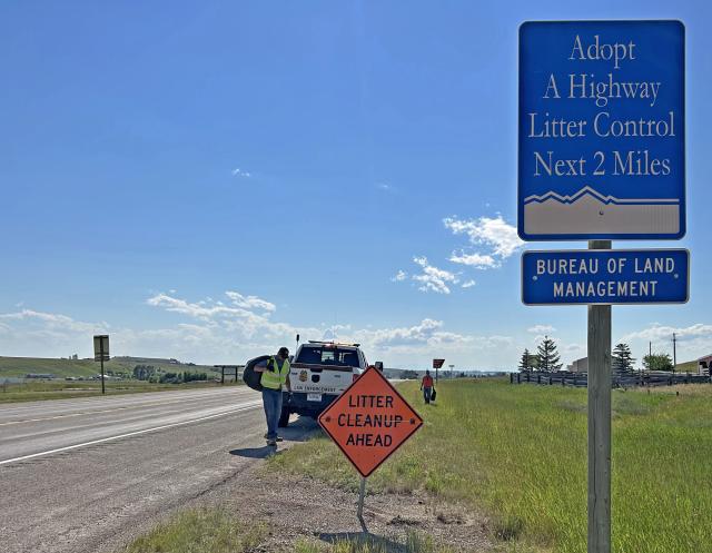 Photo of an Adopt A Highway sign designating the BLM North Central Montana District’s section of Highway 87, east of Lewistown, Montana. Orange-colored safety sign and law enforcement pickup truck in the background along the highway shoulder.