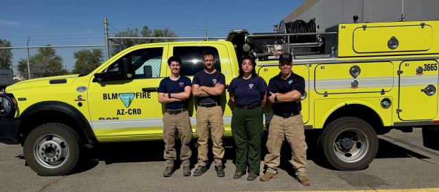 A crew of four firefighters stand in front of their fire engine.