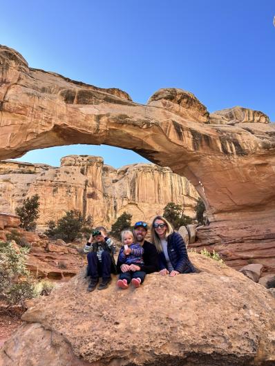 family of two adults, a man and a woman, sit with two children on a rock with an arch formation in the background