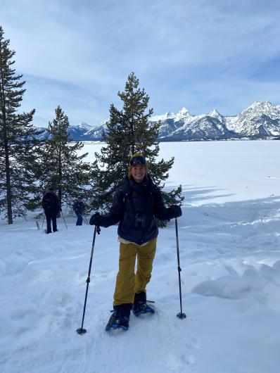 a woman poses standing in front of a conifer tree wearing snow shoes and holding hiking poles there is snow on the ground
