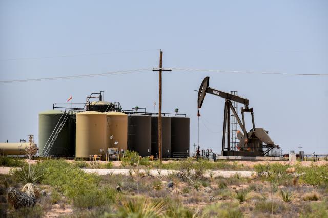 Oil pumpjack operating next to large storage tanks in a dry, desert landscape under a clear sky.