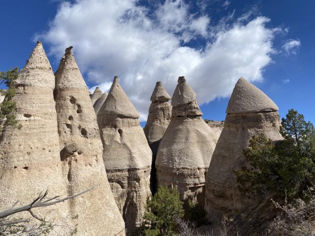 Cone-shaped rock formations, known as hoodoos, rise beneath a blue sky with scattered clouds and surrounding sparse vegetation.