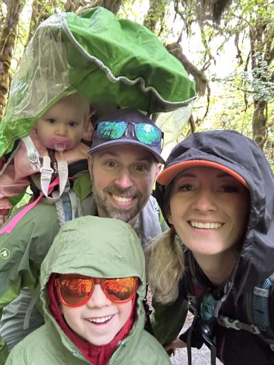 two adults, a man and a woman, the man wears a hiking backpack with a small child seated in it another small child wears sungalsses