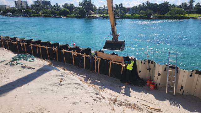 Construction workers place barriers along a sandy shoreline, while a digger overhead is ready to scoop materials by the blue water.