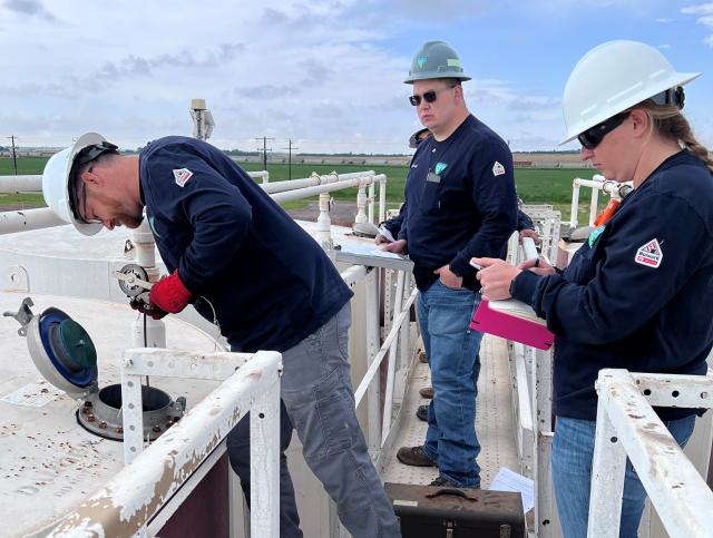 Three workers wearing hard hats and navy shirts conduct an inspection on industrial equipment outdoors with green fields in the background.