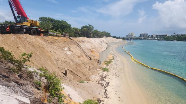 Construction site along a shoreline, featuring heavy machinery, a sand barrier, and a calm waterway lined with orange booms.