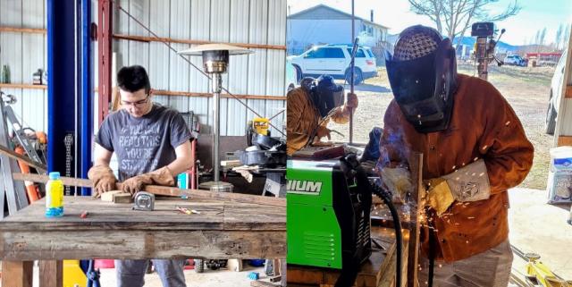 Two photos side by side. On the left, a young man wearing safety gloves works inside a shop, measuring and preparing wooden pieces for a birdhouse mount. On the right, a volunteer in protective welding gear and helmet welds the side of the metal mount, sparks flying as they work next to a green welding machine.