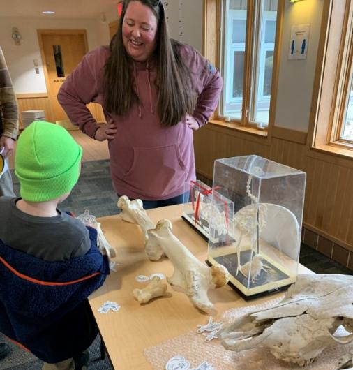 Standing around a table with exhibits of skulls and bones, an archaeologist shares information with a child in a bright green hat.