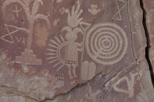 Petroglyphs carved into red/brown rock, featuring a spiral, a figure with a headdress, and geometric shapes.