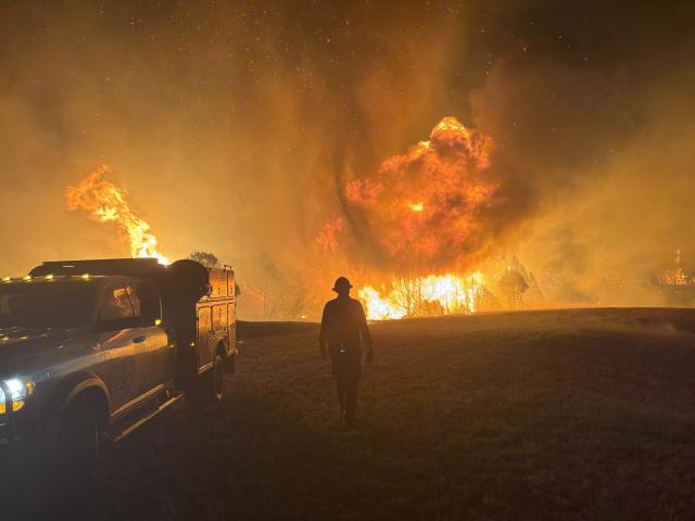 A wildland firefighter is silhouetted by a wildfire in the background as they walk near a truck.