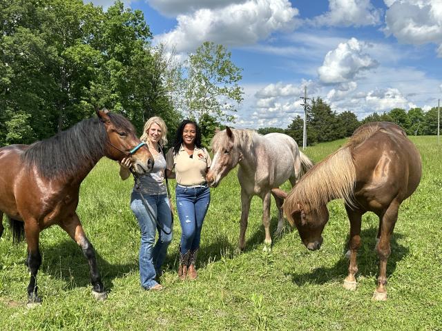 Two people stand in an open grassy field smiling with three horses.