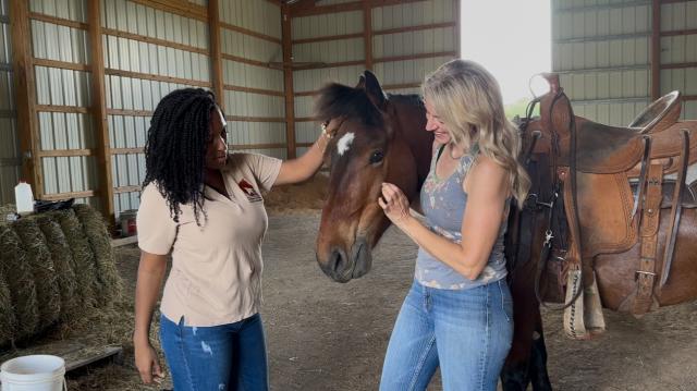 Two women gently interact with a brown horse inside a barn, one petting its mane while the other stands close by.