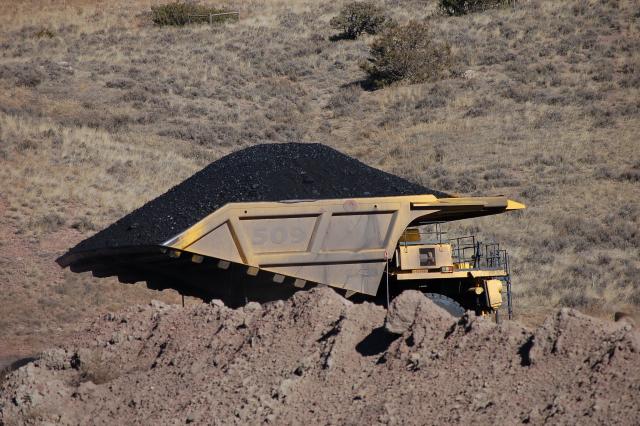 A large yellow dump truck loaded with coal sits with dirt in the foreground and a field with scrub brush in the background.