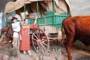 Kids in pioneer clothes stand next to covered wagon