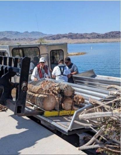 Volunteers and staff on a boat deck load brush-bundle habitats while the vessel is moored to a dock, with the lake and shoreline in the background.