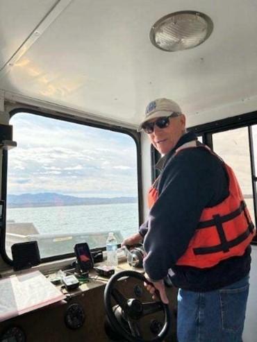 A volunteer wearing an orange life jacket steers the vessel, with the lake visible through the front window.