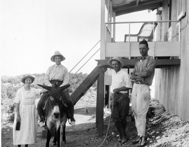 a historic photo of a group of people outside a building. Two men and two women. One woman sits on a burro.