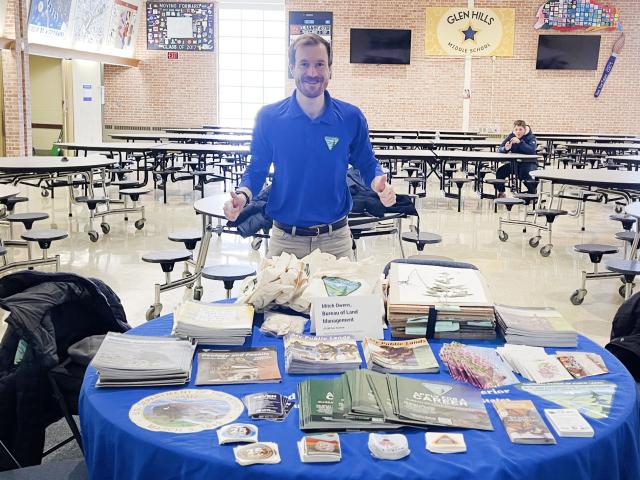 A person stands beside a table filled with brochures and materials, in a spacious school cafeteria with empty tables.