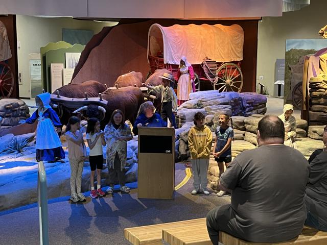 A woman stands behind a podium with children on either side of her. Behind her, is a reenactment of the Oregon rails with the wagon. 