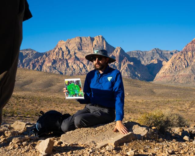 A man sits on the ground holding up a picture with hills in the background.