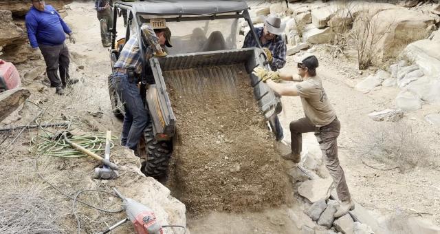 A group of three men are pushing on a dump truck that is spilling dirt onto the ground. 