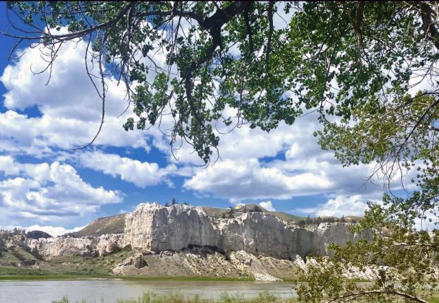 White limestone cliffs rise above a calm river under a bright blue sky with scattered clouds, framed by green leafy branches.