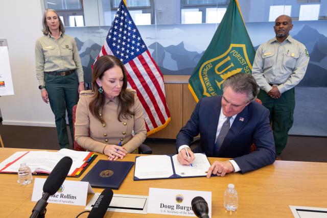 Secretary of the Interior Doug Burgum and Secretary of Agriculture Brooke L. Rollins sit together at a table signing a joint memo on wildfire preparedness with staff and standing next to flags in the background at the USDA Forest Service.