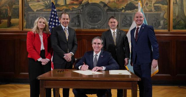 Secretary of the Interior Doug Burgum (center and sitting) signs an agreement for the Emery County Land Exchange with Michelle McConkie, Executive Director, Utah Institutional Trust Lands Administration (far left), Utah Senator John Curtis (left), Acting BLM Director Jon Raby (right), and Utah Governor Spencer Cox (far right), standing around him at the table.