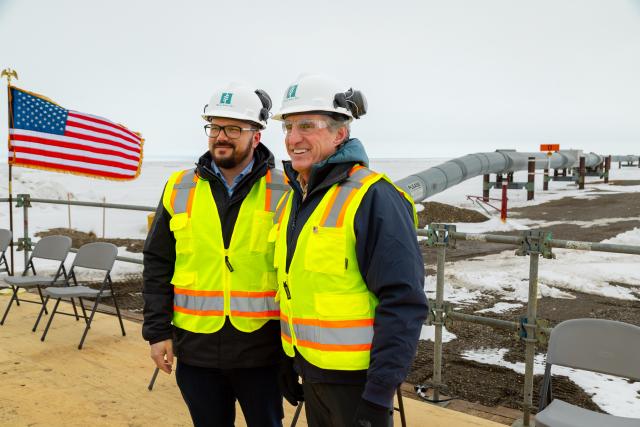 Secretary of the Interior Doug Burgum with an energy worker wearing reflective vests and hard hats in front of the Trans-Alaska-Pipeline.