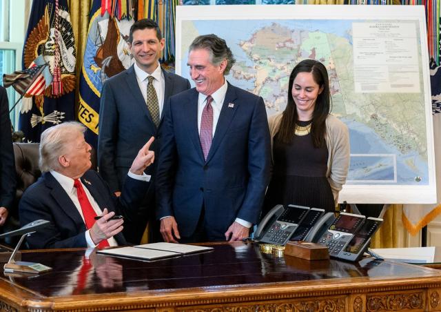 President Donald Trump sitting at his desk pointing at Secretary of the Interior Doug Burgum at the signing of an Executive Order approving the Ambler Road Project in Alaska. Both are surrounded by other staffers.