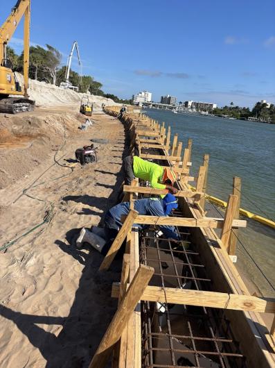 Construction workers prepare a concrete form along a beachside, with heavy machinery and a waterway in the background under sunny skies.
