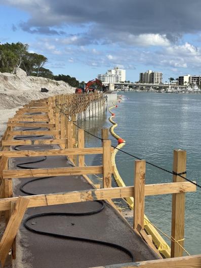 Construction site along a waterfront, featuring wooden frameworks, an excavator, and a yellow containment barrier in the water.