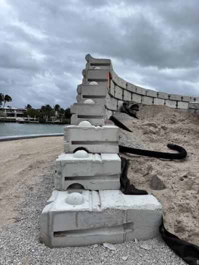 Stacked concrete barrier blocks form a retaining wall next to a mound of sand, under a cloudy sky near a waterfront.