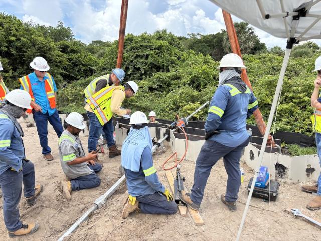 A group of workers in safety helmets and vests collaborates on-site, focusing on equipment amidst a green background.