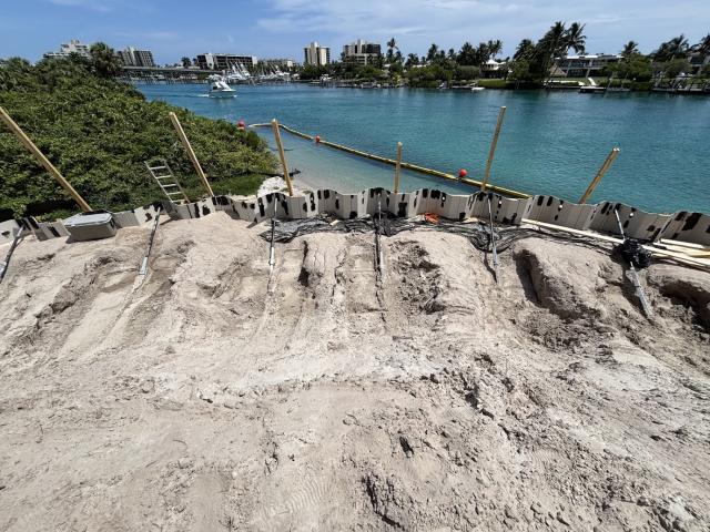 Construction site view showing sandy terrain with equipment and a waterway lined with palm trees in the background.