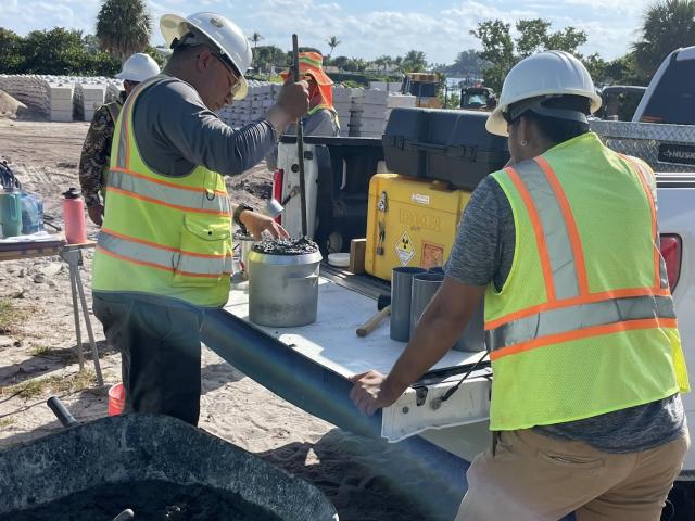 Two workers in safety vests and helmets conduct material testing on a construction site, using equipment on the tailgate of a pickup truck.