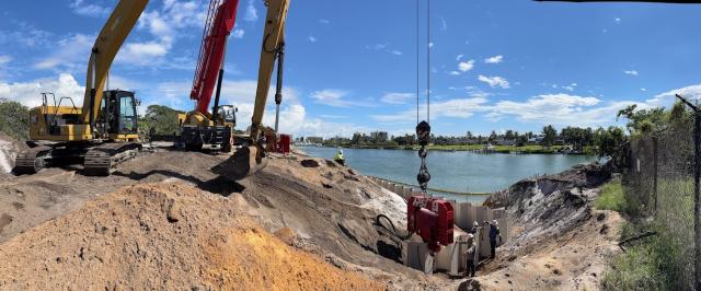 Construction site with heavy machinery near a waterfront, showcasing excavators, workers, and a clear blue sky.