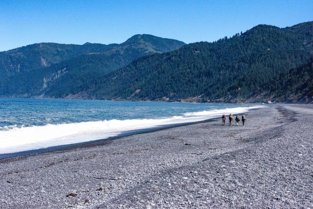 four hikers walk along beach near mountains and water on a blue bird day.