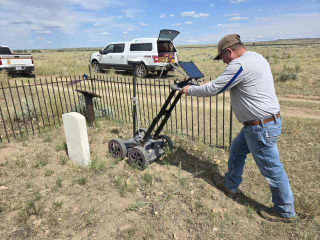 Man operating ground-penetrating radar near a fenced grave marker in a dry, grassy field under a partly cloudy sky.