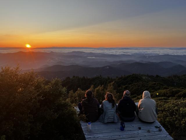 Volunteers sit on a wooden plank looking west into the singing oragne sun over the mountians and ocean.
