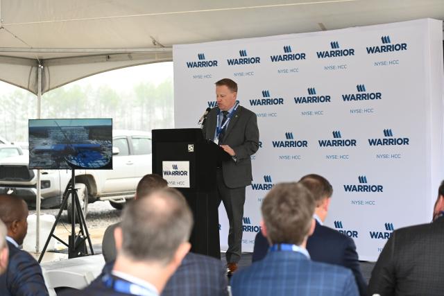 A speaker delivers a presentation at a promotional event under a tent, with a screen displaying a construction project in the background.