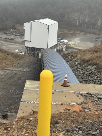 Industrial site featuring a large storage building with a curved metal roof, surrounded by gravel and construction materials. A yellow guardrail is visible.