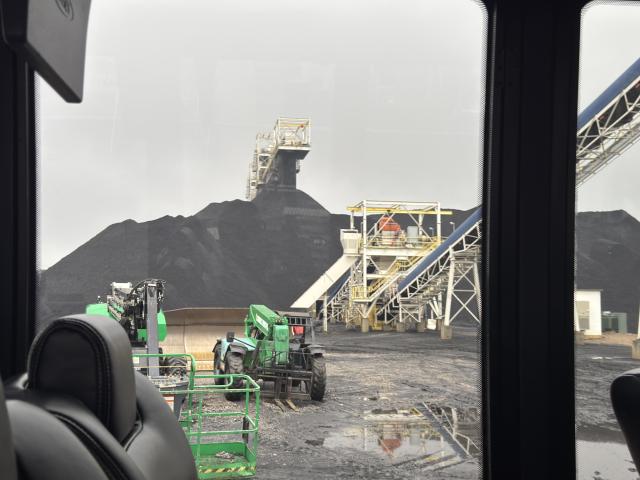 View from a vehicle showing heavy machinery and large coal piles at an industrial site, with conveyor belts and cloudy sky.