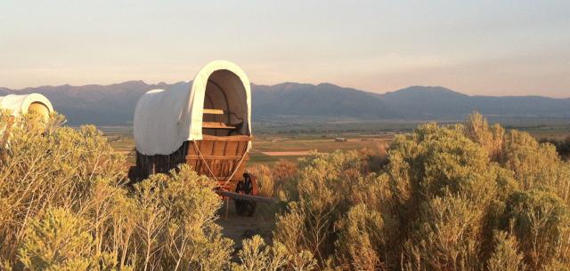 A covered wagon surrounded by sage brush with distant mountains at sunset