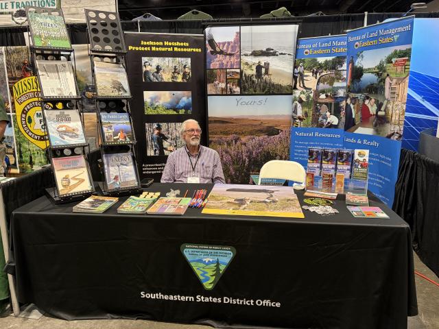 Person sitting at an expo booth surrounded by pamphlets and informational displays