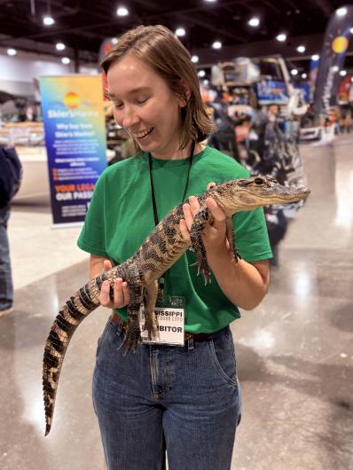 Person wearing a green shirt and jeans holding a young alligator at an indoor event with various booths in the background.