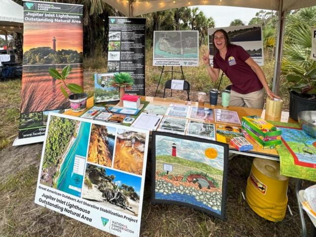 Outdoor booth under tent showcasing Jupiter Inlet Lighthouse natural area, with informational posters, maps, and a person posing with a peace sign.